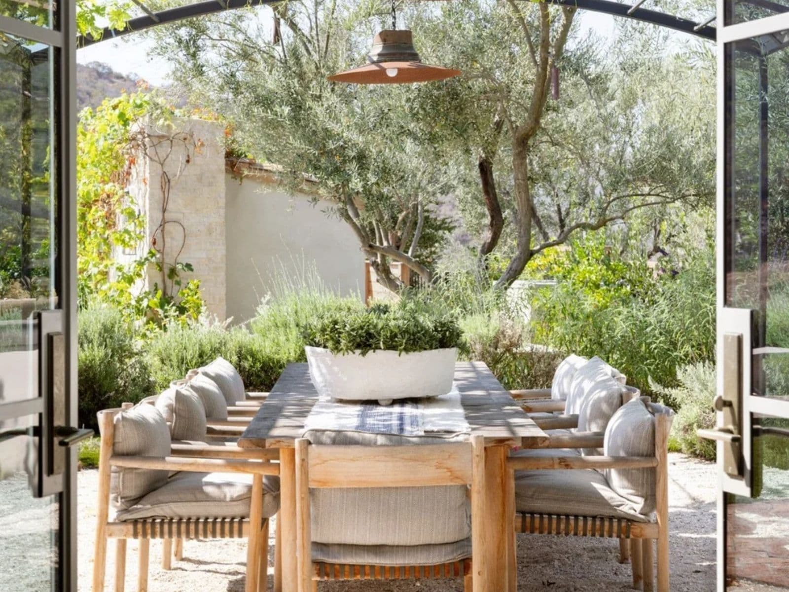 Teak dining table and armchairs on gravel under a grape arbor